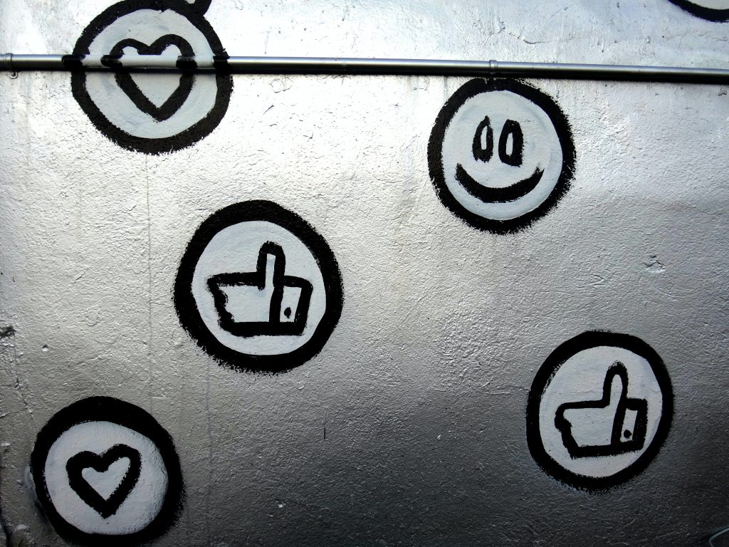 A graffitied silver wall, with black and white social media icons painted into it. This includes the Facebook like, the Facebook love react, and a smiley face.